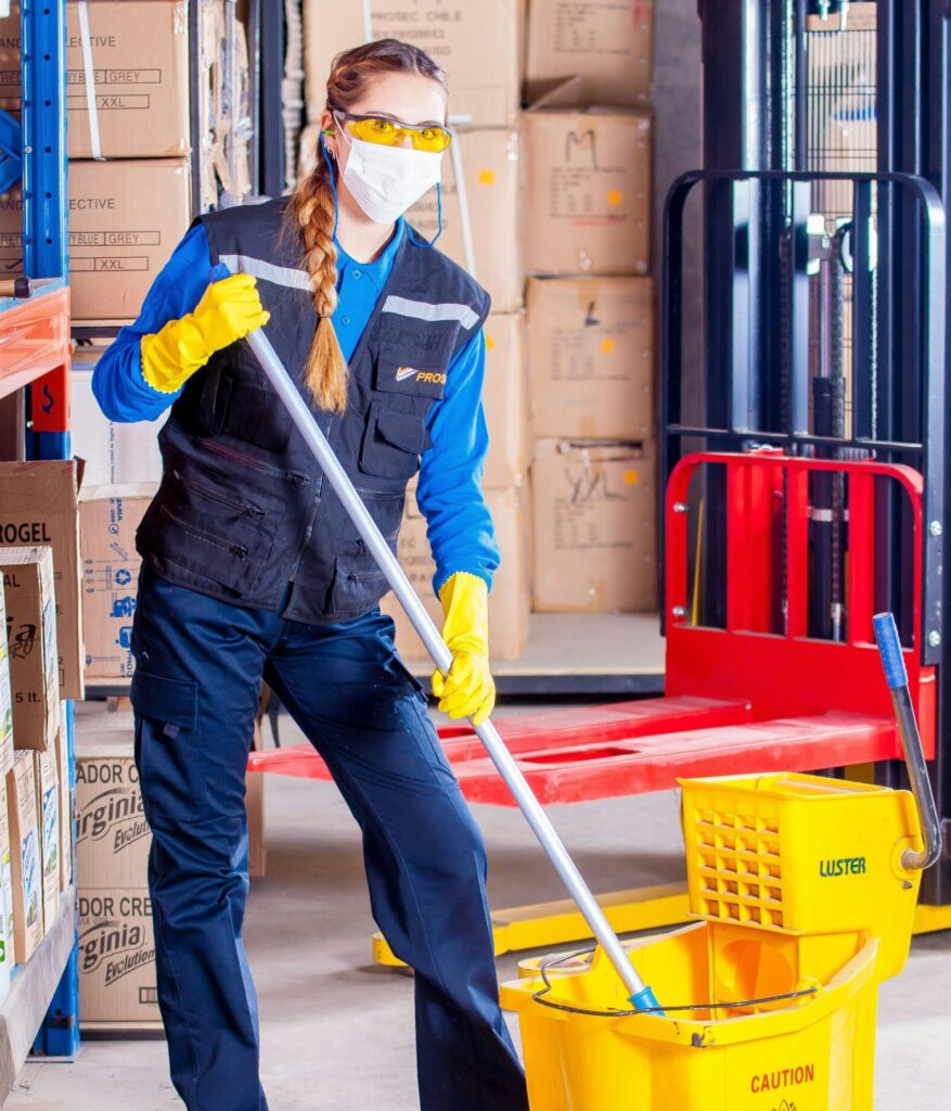 Female janitor in safety gear cleaning a warehouse floor with a mop and bucket.